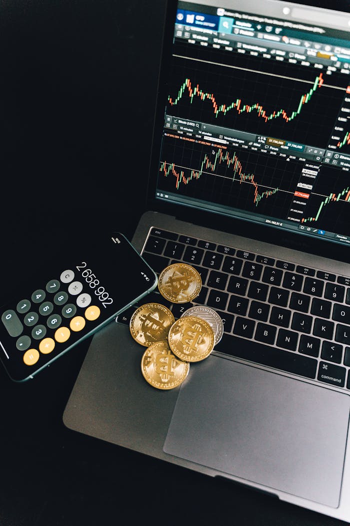 A still life of Bitcoin coins, a smartphone calculator, and a trading chart on a laptop.